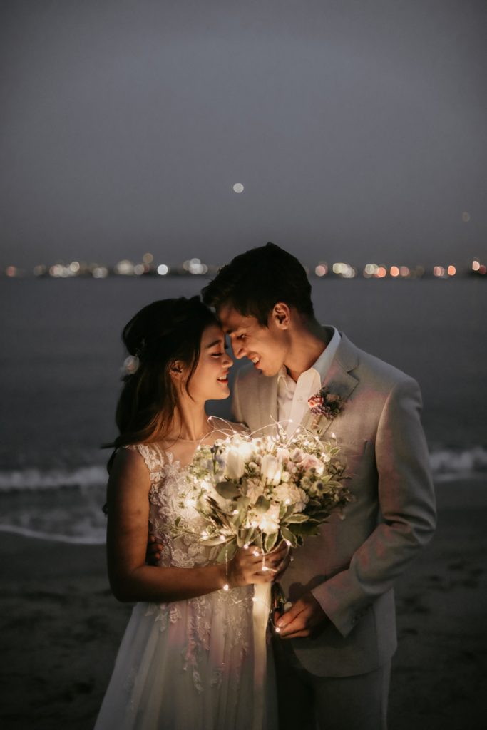 In questa foto due sposi si guardano innamorati mentre posano al crepuscolo su una spiaggia con alle spalle il mare. I loro visi sono illuminati solo dal bouquet luminoso che la sposa tiene con la mano destra