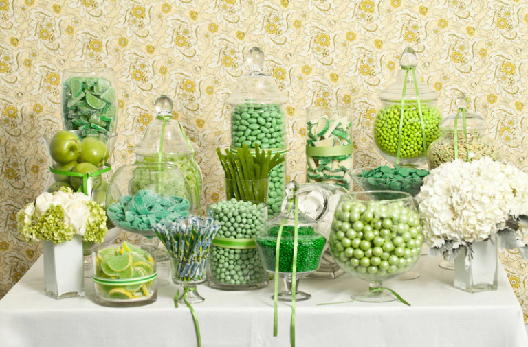 In questa foto una confettata con diversi toni di verde e bianco. Sul tavolo 14 jar di vetro, ognuna contenente diversi dolciumi: onfetti verdi color menta e color salvia, mele verdi, liquirizie verdi, caramelle verdi e spicchi di limoni. Ad adornare il tavolo, alle estremità, anche due vasi con ortensie bianche.  