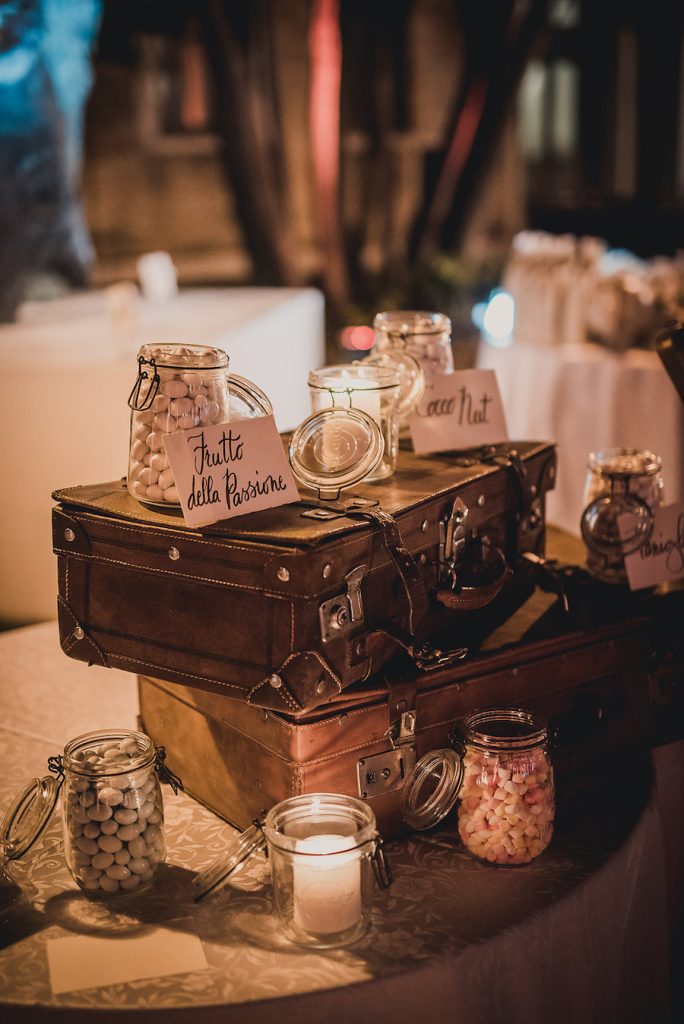 In questa foto una confettata per matrimonio a tema viaggio realizzato con valigie vintage su cui sono poggiati vasetti di vetro pieni di confetti e candele. Davanti ad ogni vasetto è appoggiato un cavaliere con i gusti dei confetti 