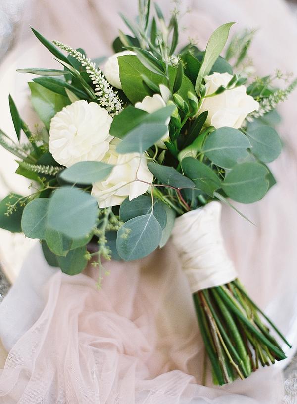 In questa foto un esempio di bouquet per la promessa di matrimonio. Un mazzo di fiori bianchi, eucalipto e rami di ulivo. Il mazzo è legato con un nastro bianco ed è adagiato su un velo rosa cipria.