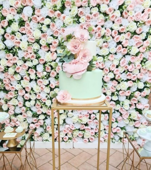 In questa foto una torta a due piani con pasta di zucchero colore verde menta chiaro, riposta su un espositore di legno. La torta è decorata con lisanthium rosa e foglie verdi. Dietro la torta, una parete di fiori con rose inglesi di colore rosa e bianco e foglie verdi. 