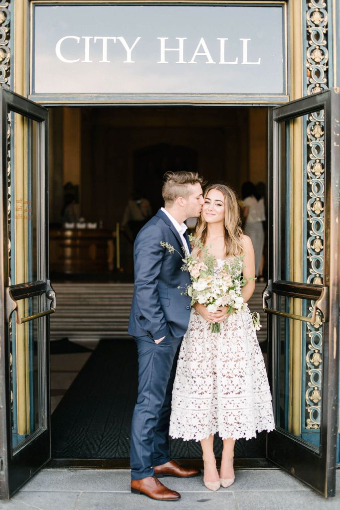 In questa foto una coppia di sposi felici davanti all'ingresso del municipio con la scritta City Hall. Lui abbraccia la sposa e le dà un bacio sulla guancia. Lei sorride e stringe tra le mani un bouquet di fiori bianchi, rosa e rametti di eucalipto ed erbe aromatiche. Lui indossa un abito blu e lei un abito bianco in pizzo macramè. 