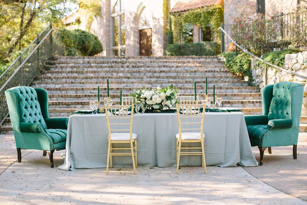 In questa foto, un tavolo allestito con una tovaglia color verde salvia e sopra un runner verde ottanio. Ad adornarlo un centrotavola con rose bianche ed eucalipto, sei candelabri dorati con candele, sempre in verde ottanio. Attorno al tavolo 4 sedie Tiffany di color oro e sedute bianche.   ai due capotavola, poltrone in velluto verde bosco.