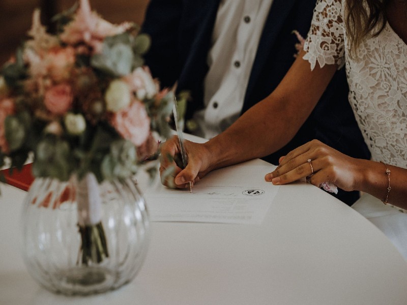 In questa foto una coppia di futuri sposi durante la promessa matrimonio al Comune. Lei firma un foglio con una penna nera. Indossa un abito bianco di pizzo. Lui è seduto alla sua destra. Indossa una giacca nera e una camicia bianca. In primo piano, sul tavolo attorno al quale è seduta la coppia, c'è un vaso con un mazzo di rose rasa, bianche e fiori di campo.