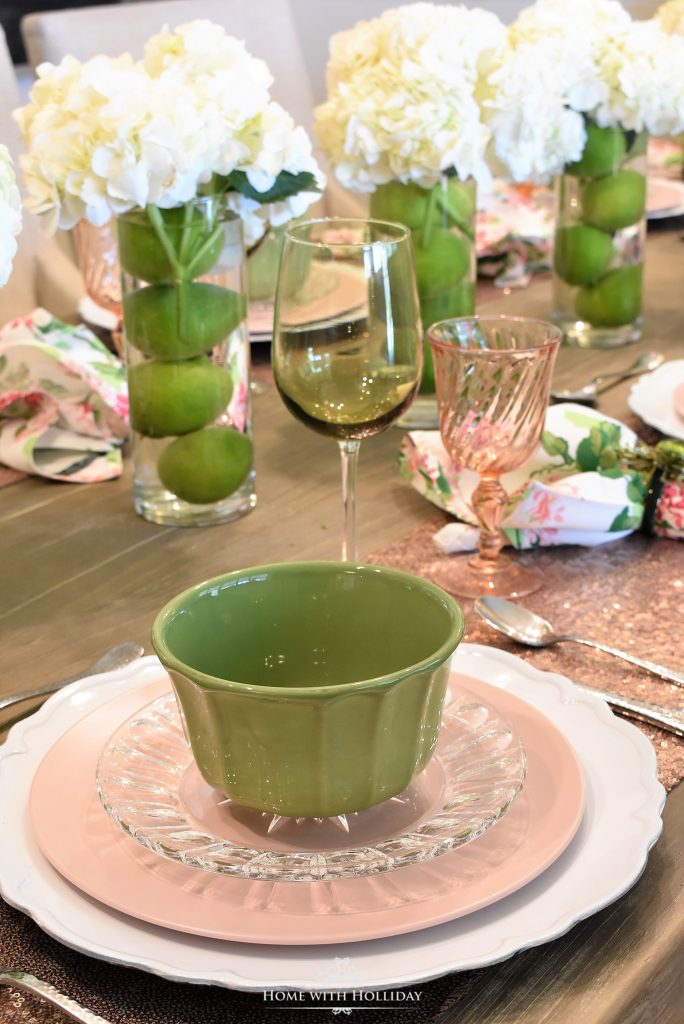 In questa foto il tavolo allestito con una mise en place dai colori verde, rosa e bianco. Sottopiatti bianchi, con sopra un piatto piano rosa e un piattino trasparente e sopra una ciotola verde. Accanto, un calice verde per il vino e un bicchiere rosa per l'acqua. Come centrotavola, tre vasi trasparenti, ognuno dei quali con 4 limoni verdi e sopra un mazzolino di ortensie bianche.  