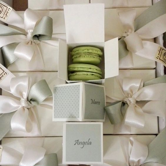 In questa foto due macaron di colore verde contenuti in piccole scatole personalizzate, con i nomi degli invitati a cui sono destinate: "Marco", "Angela". Le tre scatoline sono riposte sopra altre piccole scatole addobbate con un fiocco realizzato con un nastro di raso bianco e grigio. 