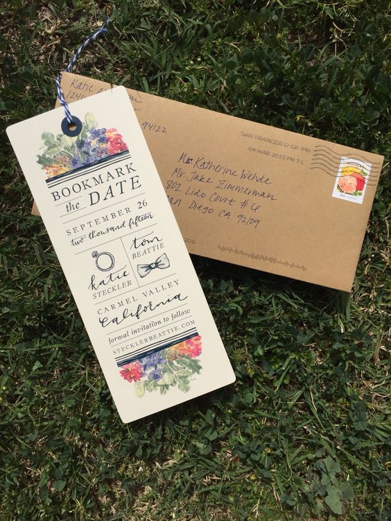 In questa foto un invito di matrimonio a forma di segnalibro abbinato ad una busta postale rettangolare di carta kraft. Sull'invito è riportata la dicitura "Bookmark the date" con i nomi degli sposi e i dettagli delle nozze. Sia sul bordo superiore che su quello inferiore sono presenti disegni floreali realizzati ad acquarello nei colori rosso, verde e blu