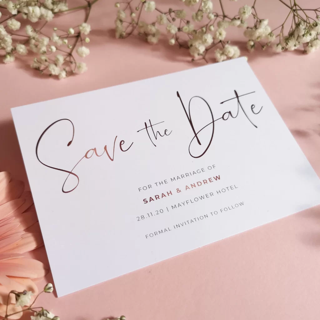 In questa foto un biglietto save the date per matrimonio bianco con scritta calligrafica grigia e oro rosa. Il biglietto è poggiato in posizione leggermente inclinata verso l'alto su un tavolo rosa ed è circondato da nebbiolina e margherite rosa