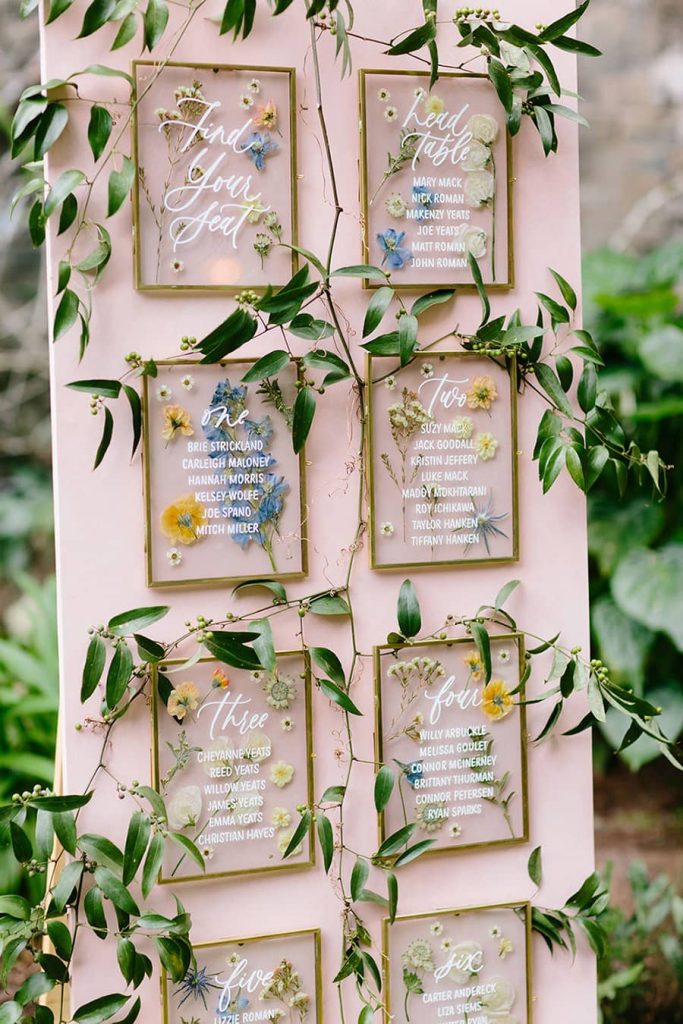 In questa foto un tableau de mariage a tema fiori realizzato con piccole cornici con vetri dipinti con fiori, il numero del tavolo e l'elenco degli ospiti. Le cornici sono appese su un rettangolo di compensato rosa cipria. Completano l'allestimento ramoscelli d'ulivo intrecciato