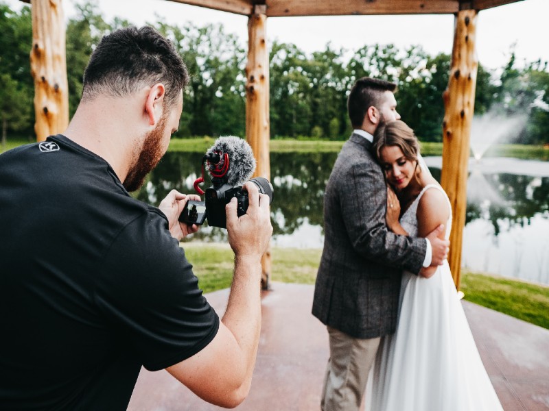 In questa foto in primo piano un operatore video matrimonio riprende una coppia di sposi abbracciati sotto un gazebo di legno. La sposa ha la testa poggiata sulla spalla dello sposo che guarda dietro di lei