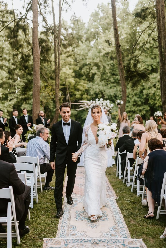 In questa foto due sposi si tengono per mano mentre  escono dalla location del loro matrimonio civile percorrendo il corridoio centrale. Il loro rito è stato celebrato in un bosco. La sposa tiene nella mano sinistra un bouquet di orchidee bianche