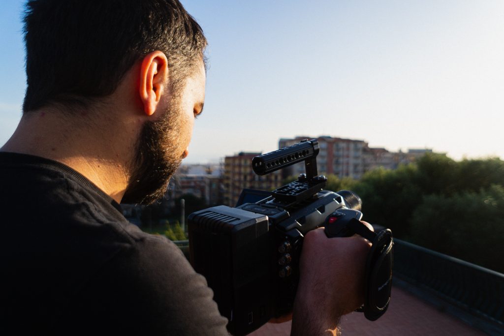 In questa foto un videomaker per matrimonio ripreso di spalle mentre tiene tra le mani la videocamera con cui gira alcune scena al tramonto