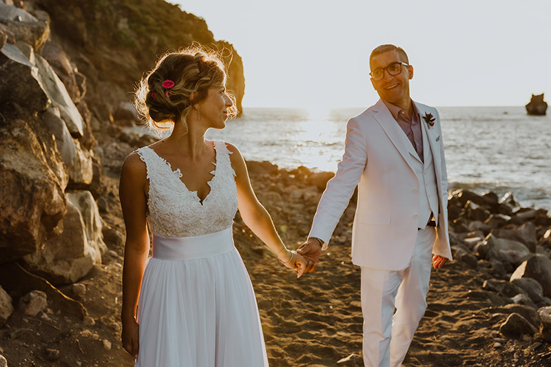 In questa foto, gli sposi si tengono per mano sulla spiaggia di Vulcano, al tramonto. Oltre alla sposa, anche lo sposo indossa uno smoking bianco e sotto una camicia grigia. 