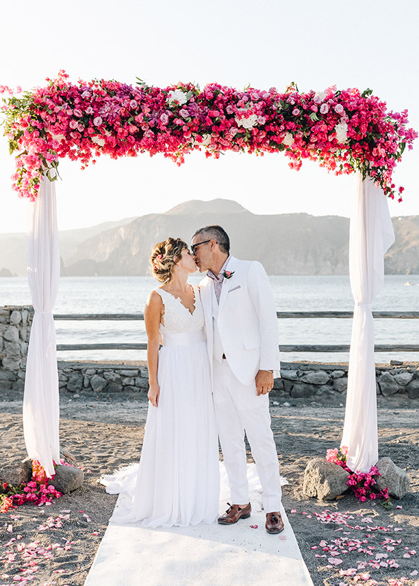 In questa foto i due sposi sotto un arco di fiori, allestito con bouganvillea fucsia. Gli sposi si baciano sotto l'arco floreale