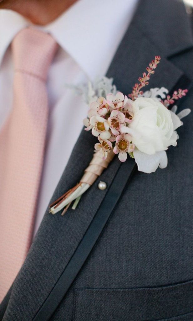 In questa foto una bottoniera con piccoli fiori rosa e marroni e una rosa bianca stretti con un nastro rosa cipria