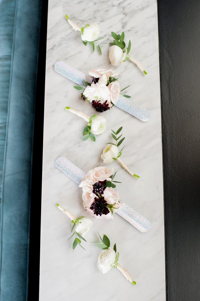 In questa foto due corsage da polso in versione Slap Bracelet con fascetta in silicone con glitter color argento poggiati su un piano di marmo. Sulla fascetta è fissata una piccola composizione di rose rosa e fiori viola. Tra i corsage sono adagiati anche 6 bottoniere composte da ranuncoli bianchi e foglioline chiusi da nastrini bianchi colore champagne