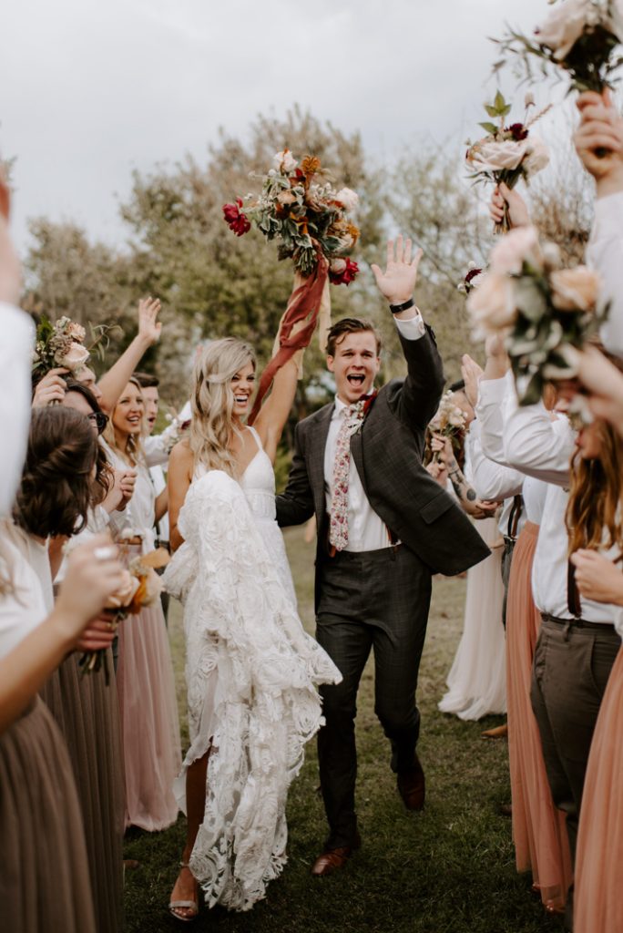 In questa foto due sposi festeggiano su un prato circondati dagli amici. La sposa tiene in alto il suo bouquet di fiori rossi, rosa e arancioni tenuti insieme da un nastro colore ruggine di lino grezzo. Lo sposo urla con la mano sinistra alzata 