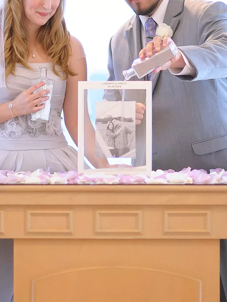 In questa foto due sposi durante il loro matrimonio simbolico celebrano il rito della sabbia versandola dentro un riquadro di vetro con all'interno una loro foto in bianco e nero. Sulla cornice del portafoto, poggiato su un tavolo ricoperto di petali di rose bianche e rosa, sono riportati i nomi degli sposi e la data delle nozze