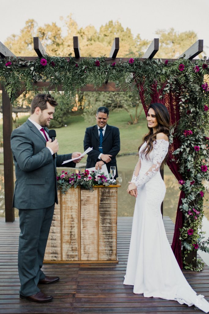 In questa foto uno sposo durante il matrimonio legge le sue promesse alla sposa che sorride. Li guarda il celebrante in piedi dietro il tavolo del rito. L'altare è addobbato con un arco di legno decorato con foglie e fiori bordeaux e un drappo di velluto rosso. Gli stessi fiori decorano il tavolo delle nozze. La sposa indossa un abito a mezza sirena con maniche in pizzo. Lo sposo indossa un abito grigio antracite e una cravatta bordeaux