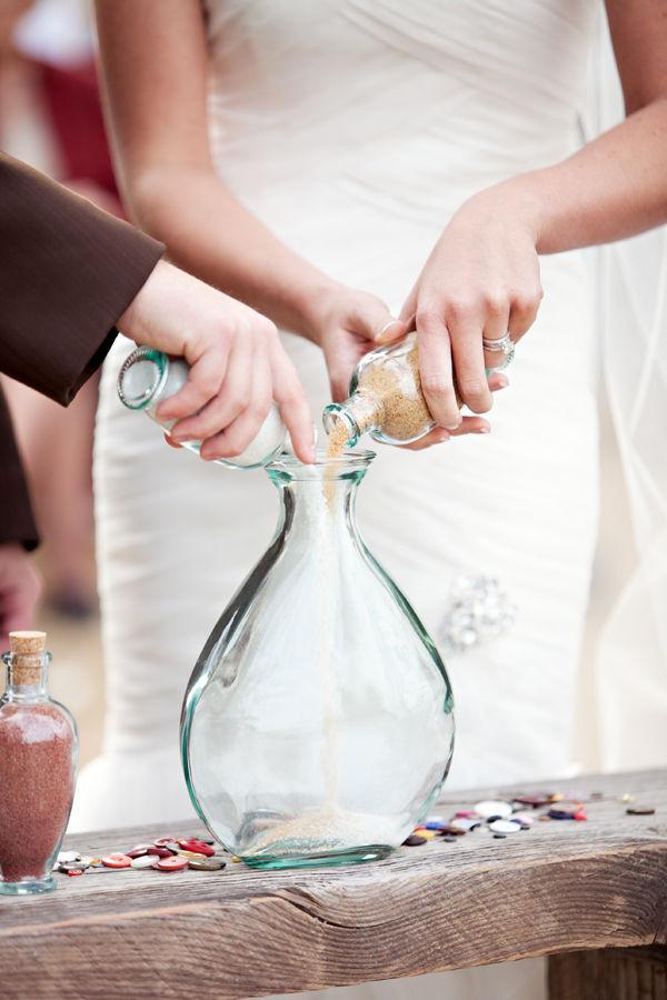 In questa foto le mani degli sposi mentre versano, durante il loro matrimonio simbolico, la sabbia bianca e naturale in un vaso di vetro poggiato su un tavolo di legno su cui sono sparsi bottoni colorati