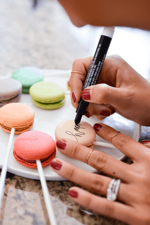 In questa foto una donna inquadrata solo sulle mani scrive su alcuni macarons colorati 