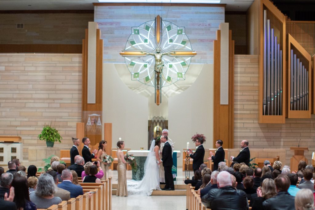 In questa foto 2 sposi si baciano all'altare di una chiesa davanti al sacerdote. A destra sono inquadrati i testimoni dello sposo, a sinistra i testimoni di nozze della sposa. La foto è un campo largo e ritrae di spalle anche tutti gli invitati seduti che assistono alla cerimonia 