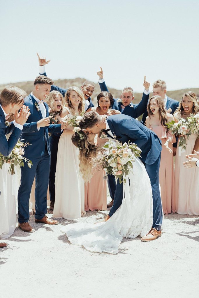 In questa foto uno sposo abbraccia la sposa facendole fare un casqué  mentre sono circondati dai loro amici e testimoni di nozze che ridono. Gli uomini indossano un abito blu navy e scarpe color testa di moro. Le donne abiti lunghi morbidi in varie gradazioni di rosa cipria. La sposa e le testimoni tengono tra le mani bouquet a fascio di fiori rosa e bianchi e foglie di eucalipto 