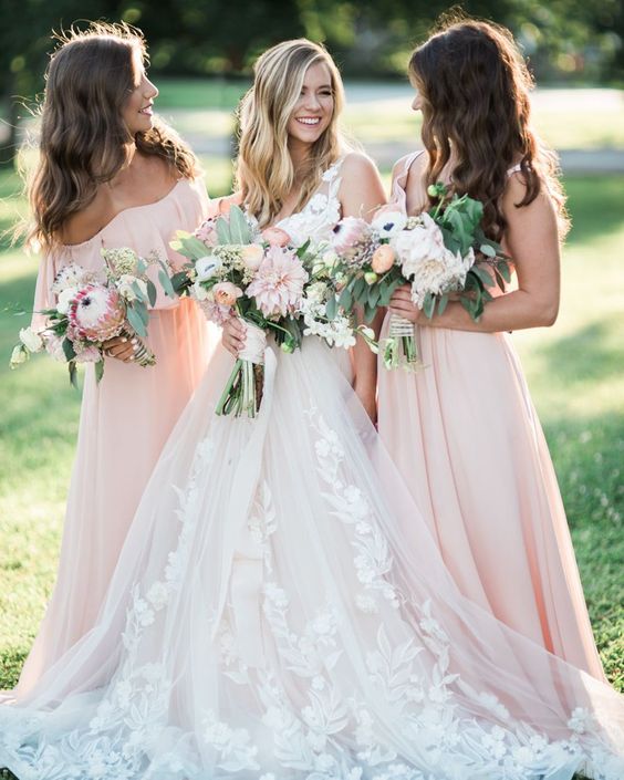 In questa foto una sposa sorride e guarda verso sinistra una delle sue testimoni di nozze che posa di profilo rivolta a destra verso di lei. Alla destra della sposa c'è la seconda testimone. La sposa tiene nella mano destra il bouquet a fascio composto di rose rosa, fiori tropicali rosa, ranuncoli di varie tonalità di rosa, anemoni bianchi e foglie di eucalipto. Le testimoni portano lo stesso bouquet ma di dimensioni più piccole. La sposa indossa un abito bianco con ramage floreale sull'orlo e su tutta la lunghezza. Le testimoni indossa un abito scivolato rosa cipria 