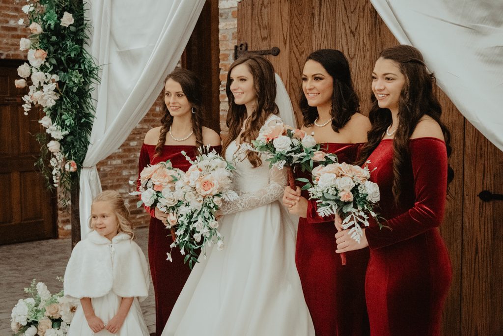 In questa foto la sposa posa con le sue testimoni di nozze e la piccola damigella, tutte rivolte verso sinistra. La sposa indossa un abito con le maniche di pizzo, il velo e porta i capelli sciolti. Tiene tra le mani un bouquet di rose rosa e bianche. Le testimoni indossano un vestito a sirena di velluto rosso con maniche e scollatura a barchetta e tengono tra le mani un bouquet uguale a quello della sposa ma di dimensioni più piccole. La piccola damigella in basso a sinistra indossa un abito bianco con una mantella di pelliccia abbinata