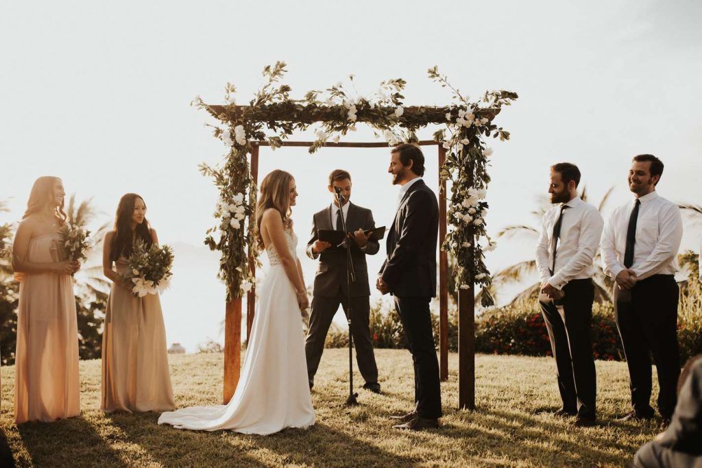 In questa foto due sposi si guardando sorridendo davanti all'altare del loro matrimonio civile creato con travi di legno e intrecciato a fiori bianchi e foglie. L'officiante legge le loro promesse di matrimonio. A sinistra sono presenti le testimoni di nozze della sposa che indossa un anito a fascia colore pesca e tengono in mano un piccolo bouquet. A destra sono presenti i testimoni dello sposo che non indossa la giacca ma solo camicia bianca e giacca