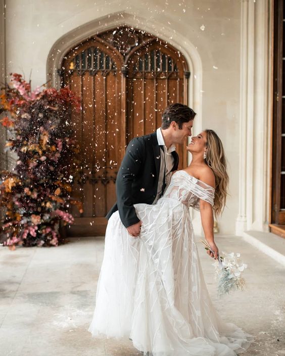 In questa foto due sposi festeggiano davanti al porto della chiesa del loro corso prematrimoniale. Lo sposo fa fare un casquet alla sposa e la guarda negli occhi. Entrambi sorridono. La sposa tiene in mano un piccollo bouquet di fiori bianchi e azzurri. Il portone della chiesa è decorato con un arco di fiori gialli, rossi, rosa e verdi