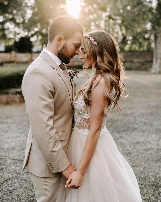 In questa foto due sposi uno di fronte all'altro si tengono per mano con le fronti appoggiate l'una all'altra, ad occhi chiusi. Lo sposo indossa un abito colore beige. La sposa una piccola corona floreale tra i capelli sciolti