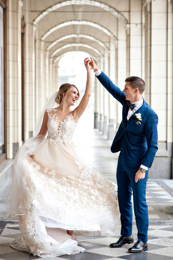 In questa foto due sposi danzano sotto un porticato. la sposa indossa un abito colore panna con decorazioni floreali sull'orlo della gonna e sul corpetto, i capelli raccolti e un velo a taglio vivo. Lo sposo indossa un abito blu navy con papillon