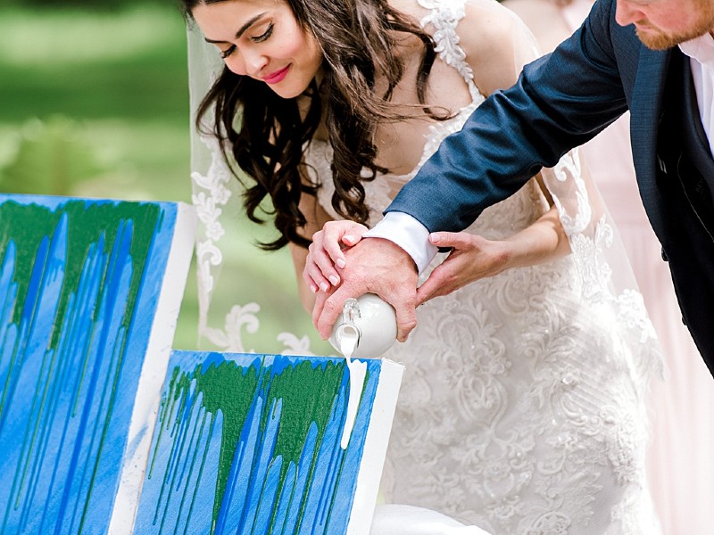 In questa foto due sposi celebrano il rito simbolico dei colori durante il loro matrimonio simbolico. Sono inquadrati il viso della sposa e il braccio dello sposo mentre versano con un bottiglietta vernice bianca su una tela già dipinta di verde e azzurro