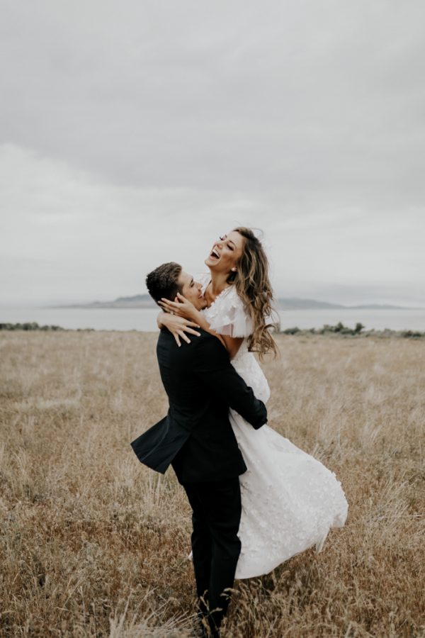 In questa foto due sposi festeggiando felici su un prato dopo il loro processetto. Lo sposo solleva la sposa abbracciandola mentre le sorride divertita