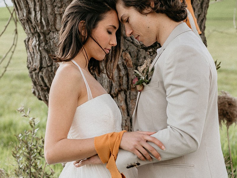 In questa foto due giovani sposi durante uno dei riti simbolici per matrimonio si trovano in piedi, l'uno di fronte all'altra, davanti ad un albero. Hanno gli occhi chiusi e le teste rivolte verso il basso con le fronti appoggiate. Lo sposo le cinge i fianchi, mentre la sposa tiene le mani sulle sue braccia. Le braccia di entrambi sono legate da un nastro di cotone colore giallo senape per celebrate il rito dell'Handfasting