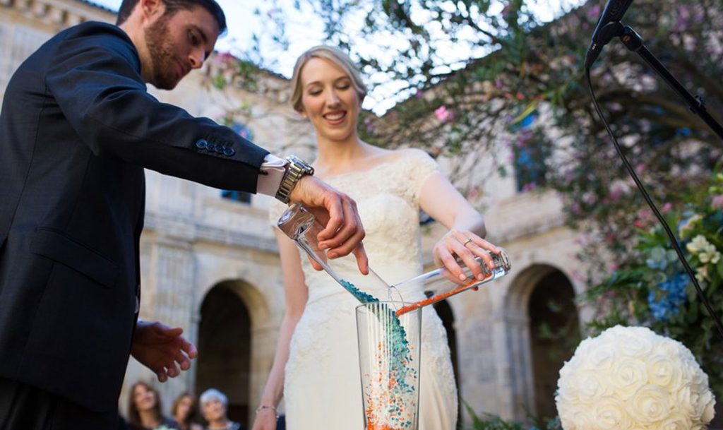 In questa foto due sposi versano dei sassolini in un vaso di vetro durante il rito simbolico del loro matrimonio. La sposa sorridente e ripresa frontalmente versa  sassolini di colore arancione con un cilindro di vetro. Con un cilindro uguale lo sposo, ripreso di profilo, versa sassolini azzurri. Alle loro spalle, sfocati, si intravedono gli ospiti 