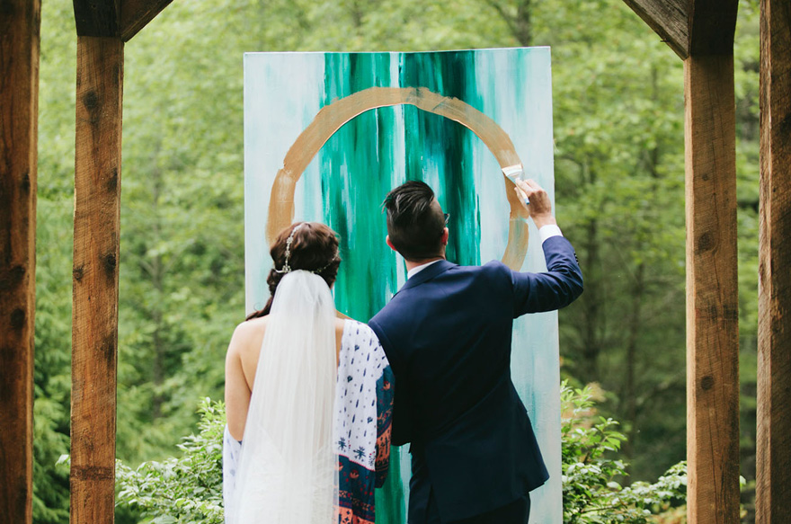 In questa foto due sposi di spalle mentre dipingono una tela durante il rito simbolico dei colori. La tela è appesa ad una trave di legno del gazebo in cui si celebrano le nozze. Sulla tela hanno dipinto due strisce verdi verticali e un cerchio dorato sopra