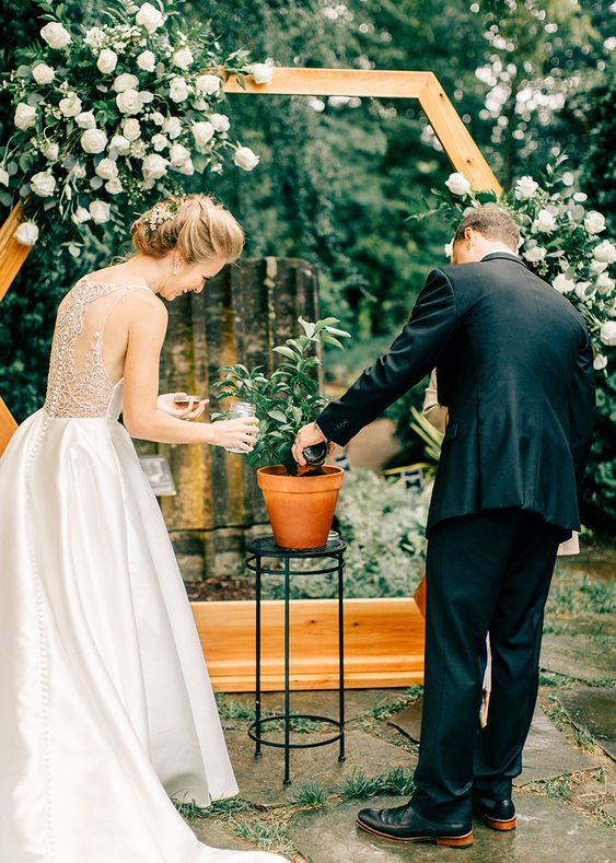 In questa foto due sposi durante uno dei riti simbolici per matrimonio particolari: il Tree Planting. Sono ripresi di spalle mentre piantano e innaffiano un piccolo albero in un vaso poggiato su un piedistallo in ferro nero. Davanti a loro è presente un altare in legno a forma di cerchio decorato con rose bianche e foglie. La sposa indossa un abito ampio in mikado e scollatura illusion sulla schiena 