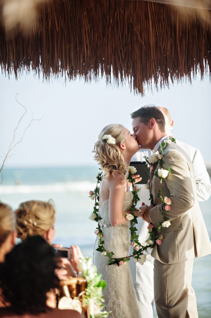 In questa foto due sposi si baciano sulla spiaggia dopo la celebrazione di uno dei riti simbolici per matrimonio tradizionali: il rito del lazo floreale. Al collo hanno legato una corda di rose bianche e rosa e di foglie. In primo piano, sull'angolo sinistro, si intravedono alcuni ospiti sfocati che che fotografano la scena 