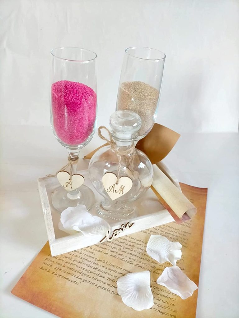 In questa foto un kit per il rito simbolico della sabbia composto da due flute, uno con sabbia rosa e uno con sabbia beige, e un vaso di vetro a forma di cuore con tappo. Legato ad ogni flute è legato un cuore di legno con le iniziale della sposa e dello sposo; legato al vaso è legato un altro cuore di legno con entrambe le iniziali degli sposi. I flute e il vaso di vetro sono poggiati su un vassoio di legno con una scritta Love sulla parte frontale