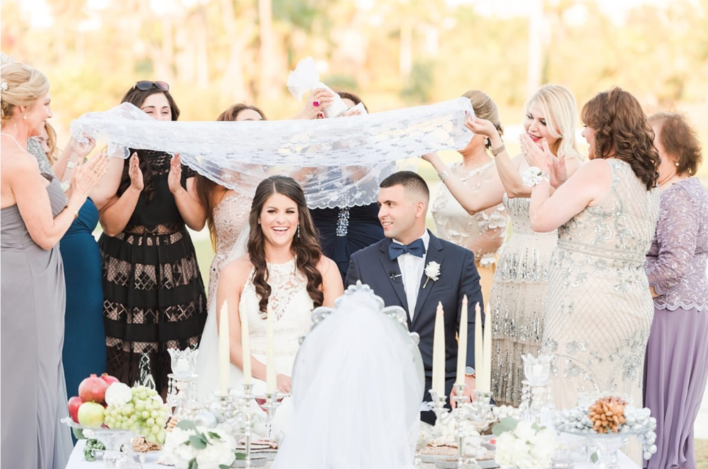 In questa foto uno dei riti simbolici per matrimonio antichi: il rituale dello zucchero. Sono ripresi due sposi sorridenti sotto ad un velo di pizzo tenuto da amici e parenti mentre una delle mamme sfrega due coni di zucchero su di loro 