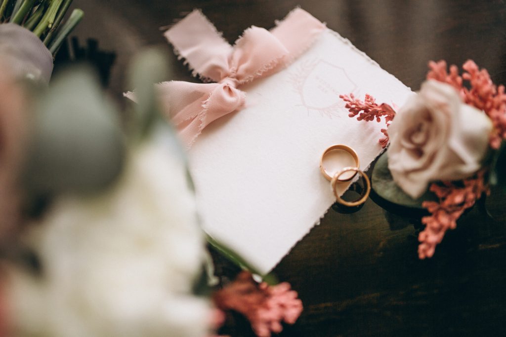 In questa foto una partecipazione di nozze in stile romantico con un nastro di stoffa chiuso con un fiocco, circondata da fiori e sulla quale sono poggiate le fedi nuziali
