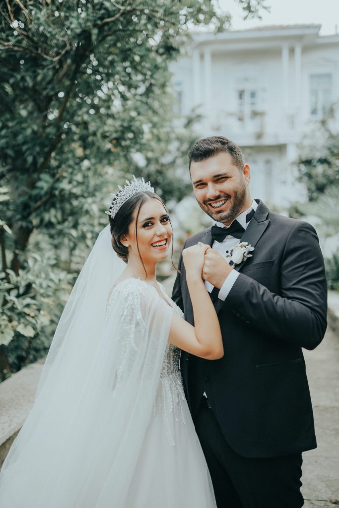 In questa foto due sposi sorridono dopo il loro matrimonio e la loro lista nozze. Sono abbracciati in un giardino e lo sposo le stringe la mano destra portandola in alto. Entrambi guardano verso il fotografo. La sposa indossa un diadema a cui è legato il velo 