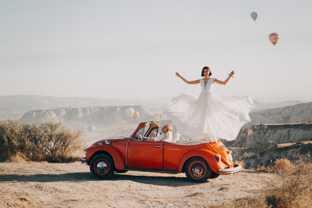 In questa foto una sposa, in piedi sulla parte posteriore di un maggiolone decappottabile arancione, fa svolazzare il suo abito mentre lo sposo la guarda seduto al posto di guida. L'auto è parcheggiata su un canyon mentre alcune mongolfiere volteggiano sullo sfondo 