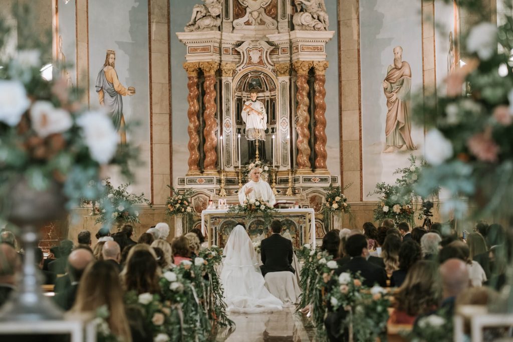 In questa foto due sposi seduti all'altare ripresi di spalle dal fondo della chiesa mentre il sacerdote recita la sua omelia. La navata centrale della chiesa è decorata con tralci di foglie e fiori rosa e bianchi