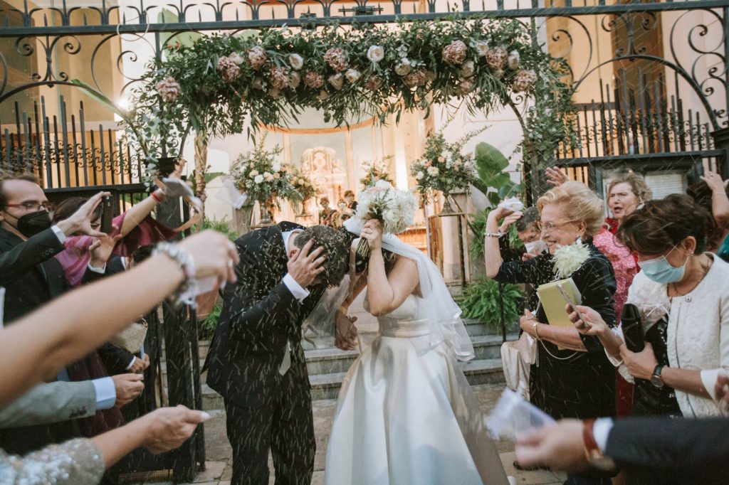 In questa foto due sposi escono dalla chiesa mentre gli invitati lanciano il riso. L'ingresso della chiesa è decorato con tralci di foglie e fiori rosa e bianchi 