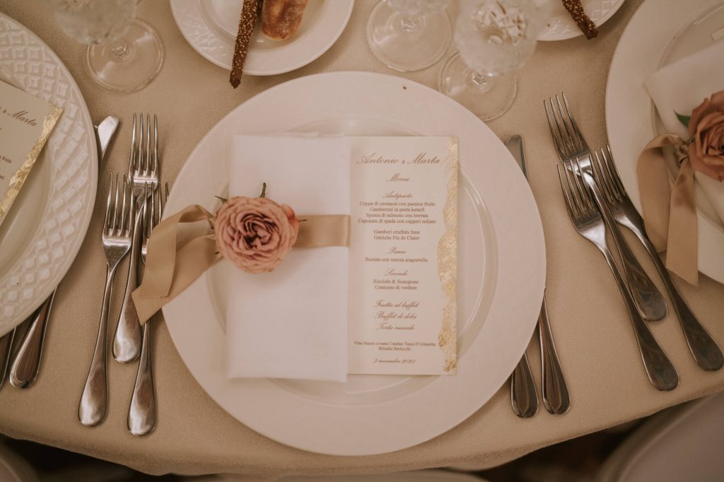 In questa foto un menù sotto ad un tovagliolo fermato da un nastro colore oro e una rosa cappuccino