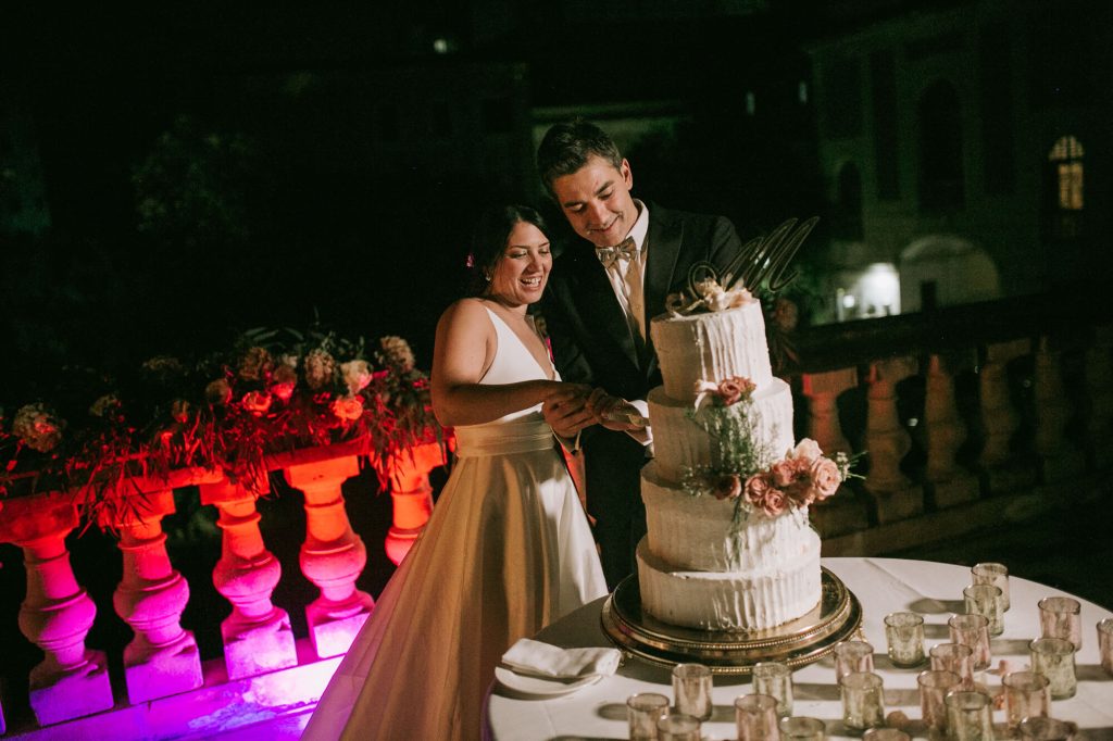 In questa foto due sposi tagliano la loro torta nuziale a piani su una terrazza illuminata 