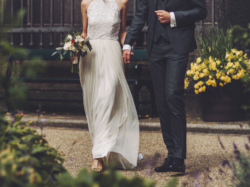 In questa foto due sposi inquadrati dal collo in giù tra i fiori camminano mano nella mano dopo le loro pubblicazioni di matrimonio in comune. Lei tiene nella mano destra un piccolo bouquet di fiori e indossa un abito scivolato. Lui un abito blu coordinato